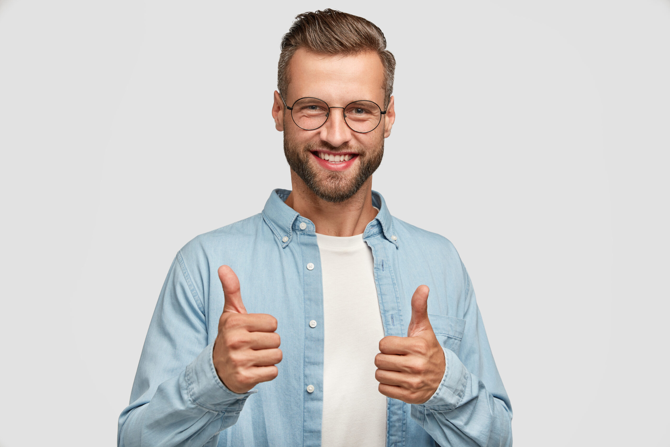 Handsome young Caucasian bearded man does okay symbol, keeps thumb raised, approves good idea of companion, has cheerful expression, stands alone agaisnt white background. Body language concept